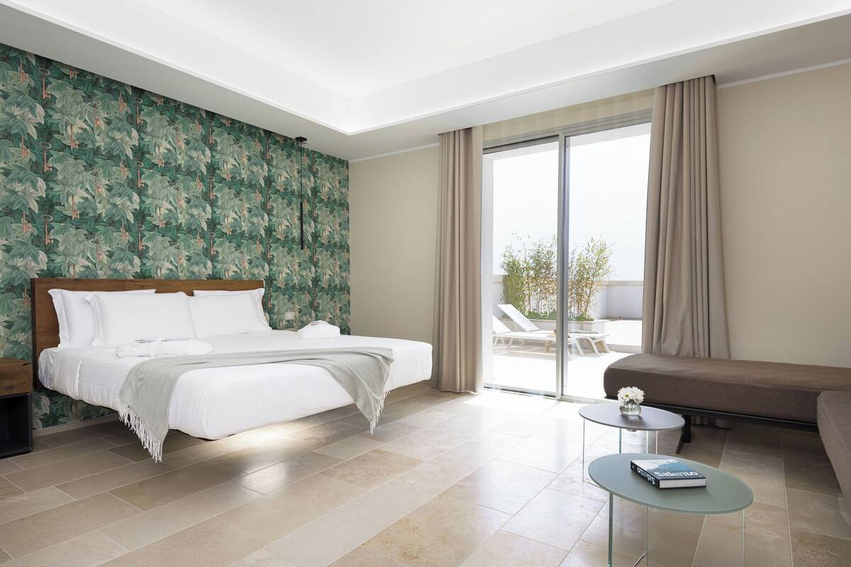 Una habitación de hotel moderna con una cama grande, una mesita de centro de cristal, un sofá y un balcón con vistas.
