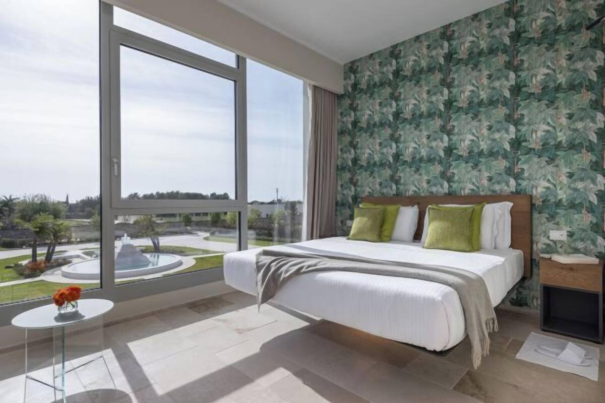 Un dormitorio con un ventanal grande, una cama con almohadas, una mesita con un florero y una pared con diseño.