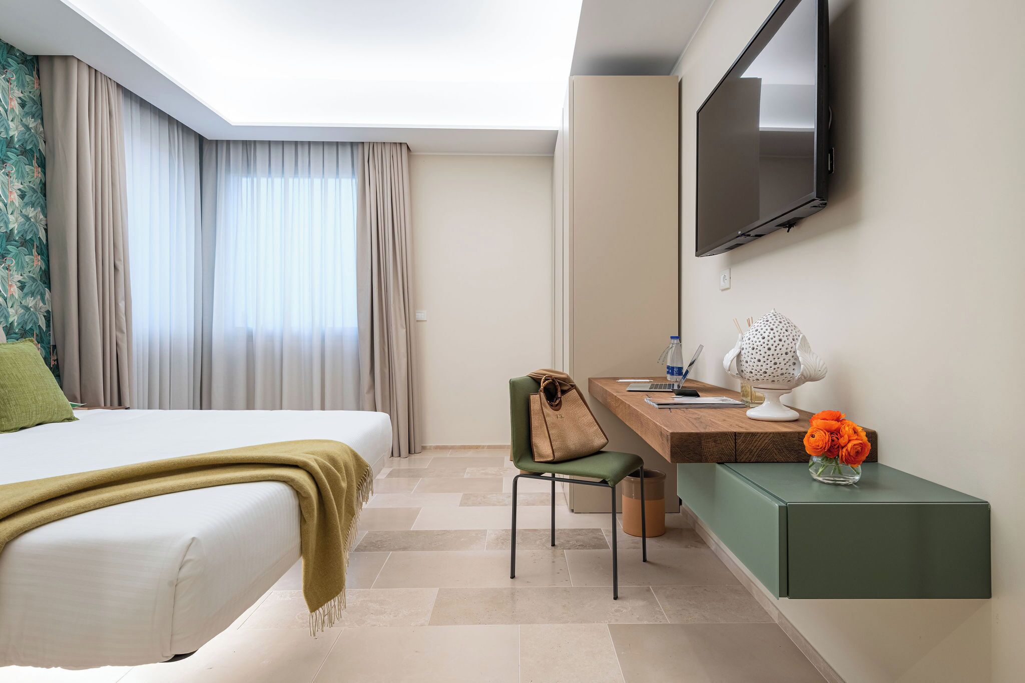 A hotel room with a bed, a desk, a chair, a television, and a vase with flowers.