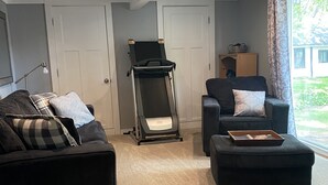 Un salón con un sofá oscuro, un sillón reclinable, una cinta de correr y un amplio ventanal con cortinas.
