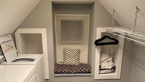 Un lavadero con lavadora, una ventana pequeña con persianas, un tendedero y un estante con ropa doblada.