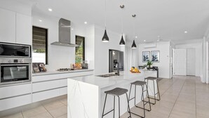 Une cuisine moderne dotée d’un grand îlot central, d’appareils électroménagers en acier inoxydable, de luminaires suspendus et offrant une vue sur l’extérieur.