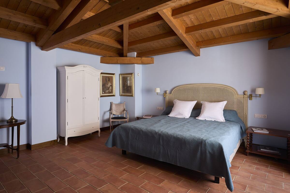 A bedroom with a bed, wooden ceiling beams, a wardrobe, and a chair.