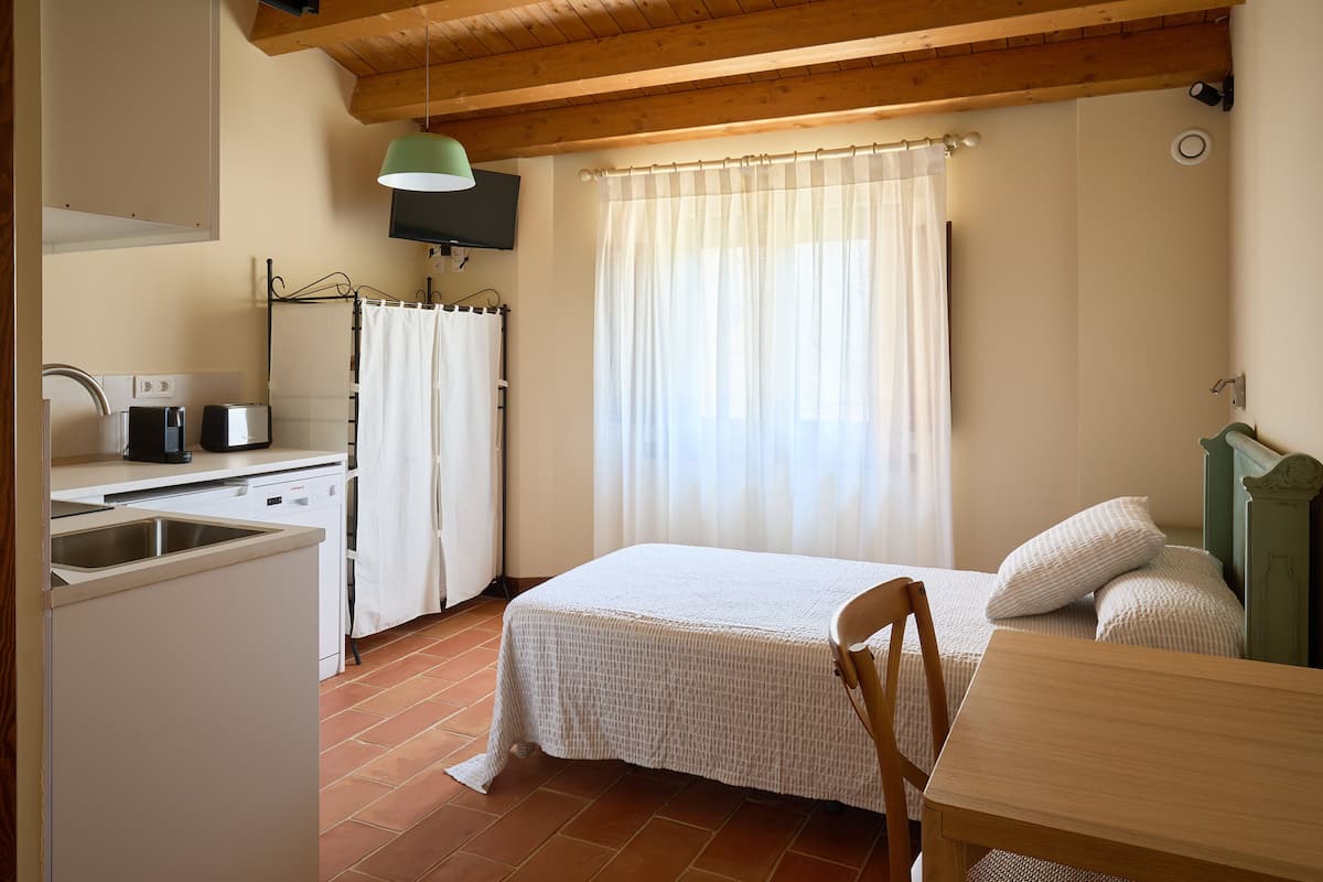 A small, cozy room with a bed, a table, a chair, a sink, a mirror, and a television.