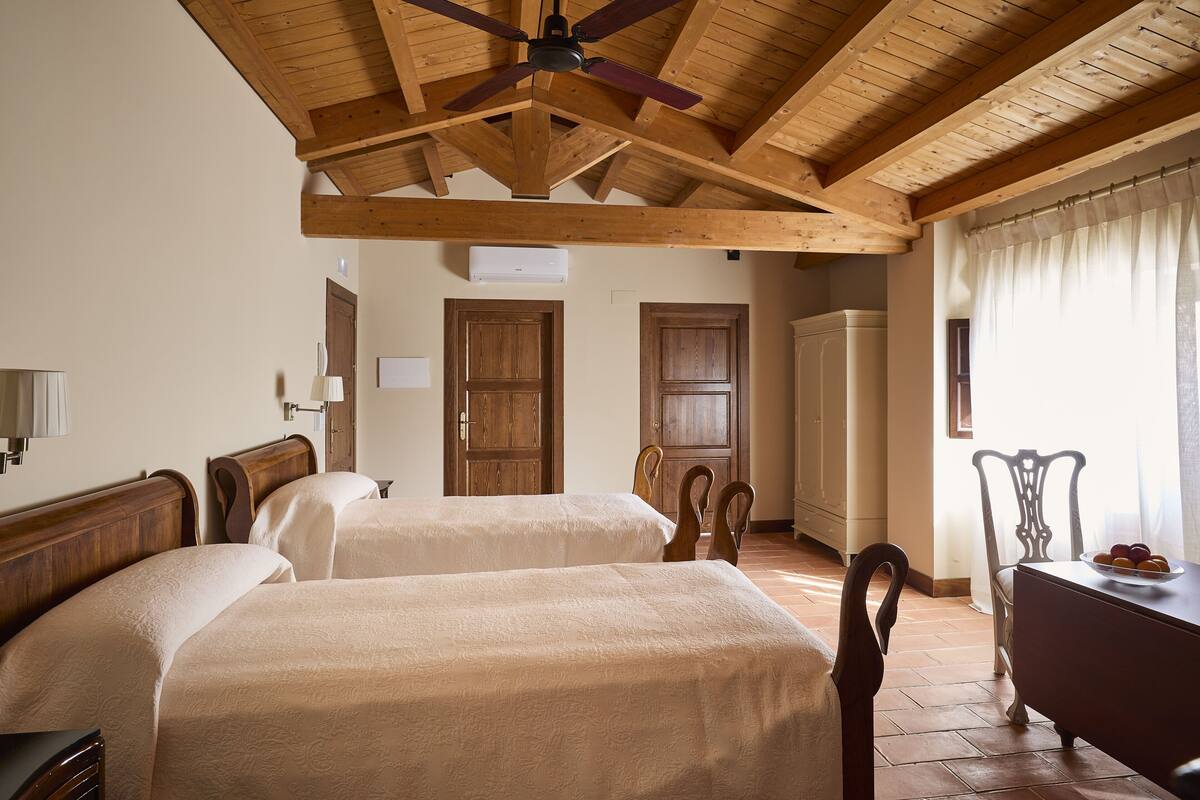 A room with two beds, wooden ceiling beams, a ceiling fan, a wooden dresser, and a window with curtains.