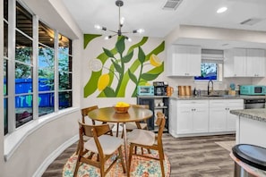 A modern kitchen with white cabinets, a dining area with a round table and chairs, and a wall mural of green leaves and yellow fruits.