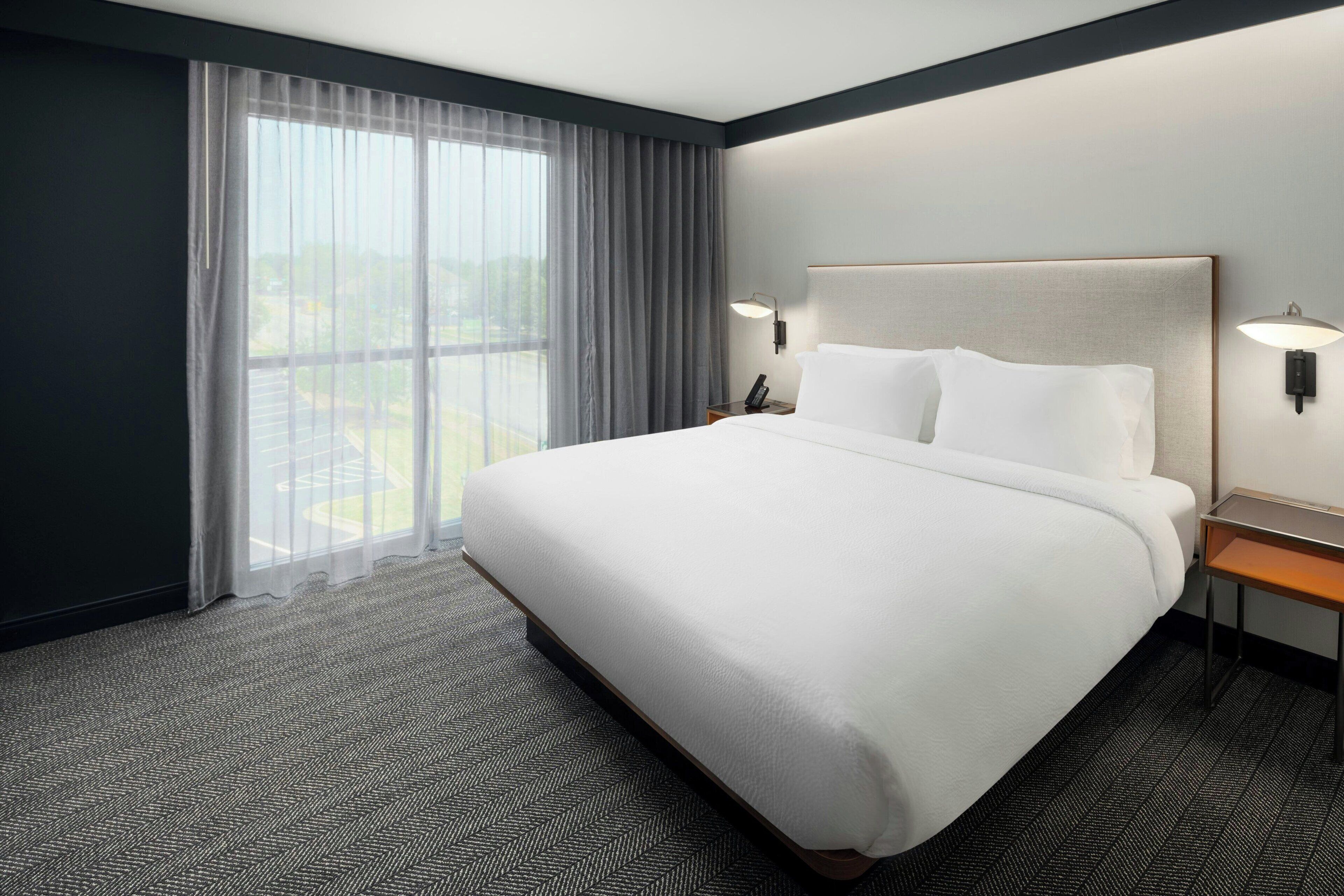 A hotel room with a large bed, a nightstand with a lamp, and a view of the outdoors through sheer curtains.