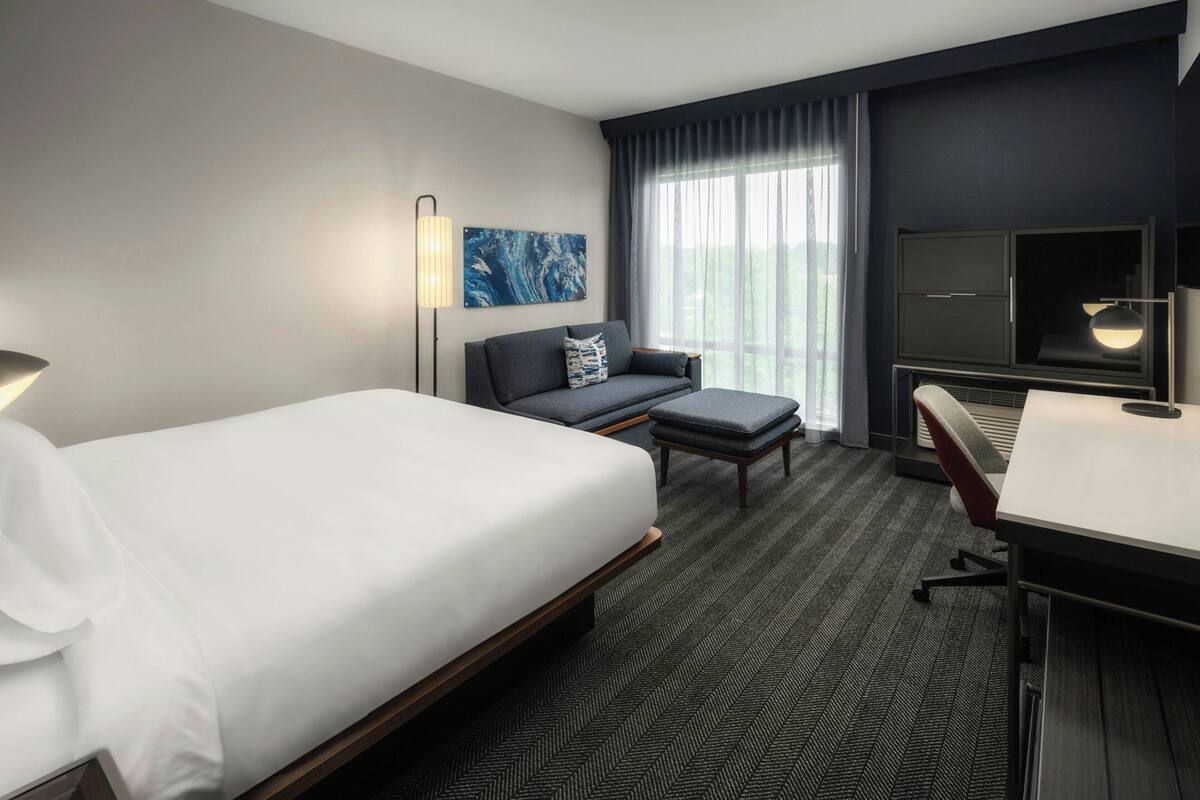 A hotel room with a large bed, a desk, a sofa, a TV, and a window with curtains.