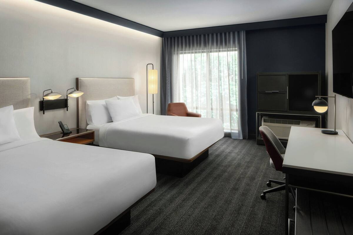 A hotel room with two beds, a desk, a chair, a TV, and a window with curtains.