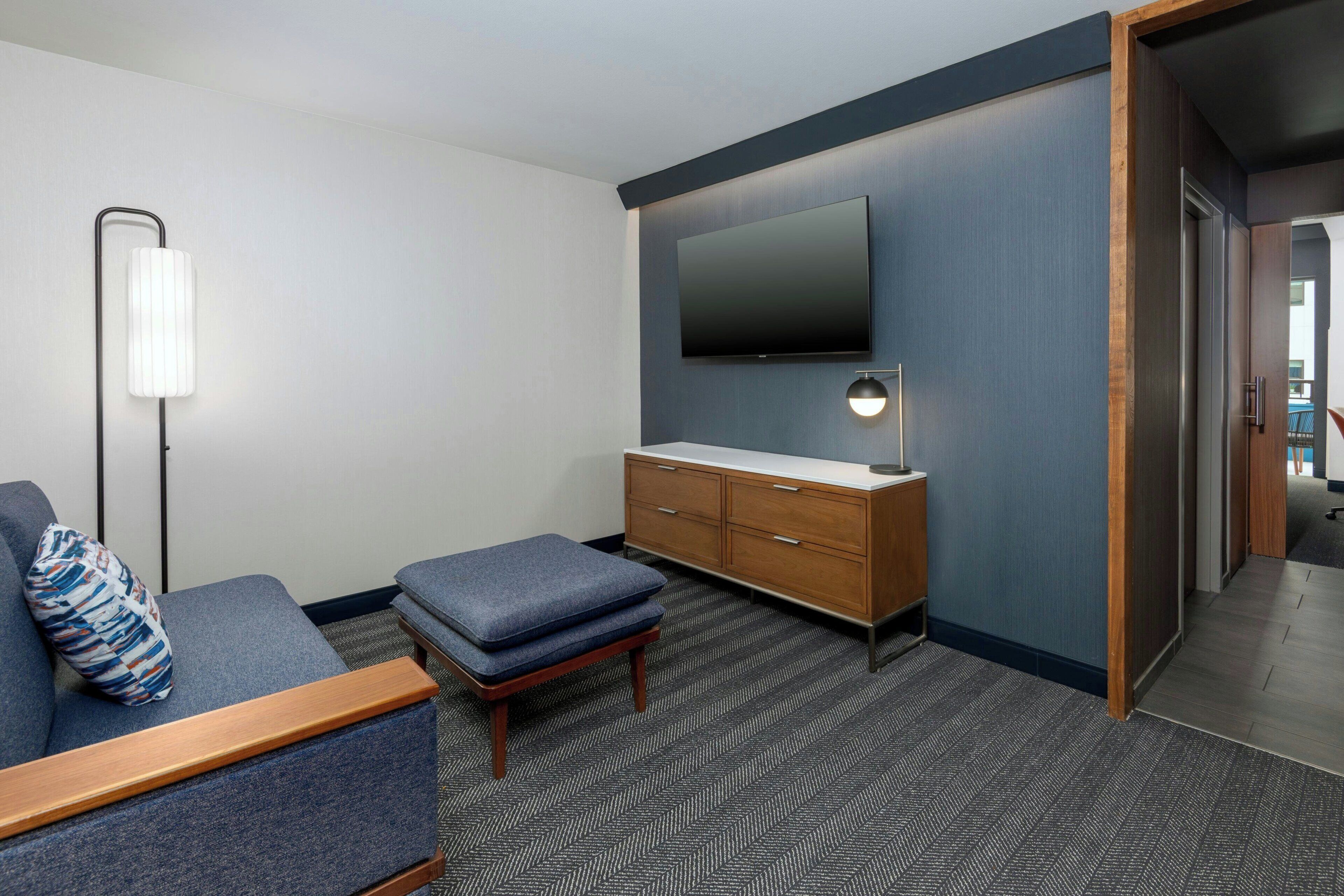A modern hotel room with a grey sofa, a wooden bench, a small table, a TV mounted on the wall, and a wooden cabinet with drawers.