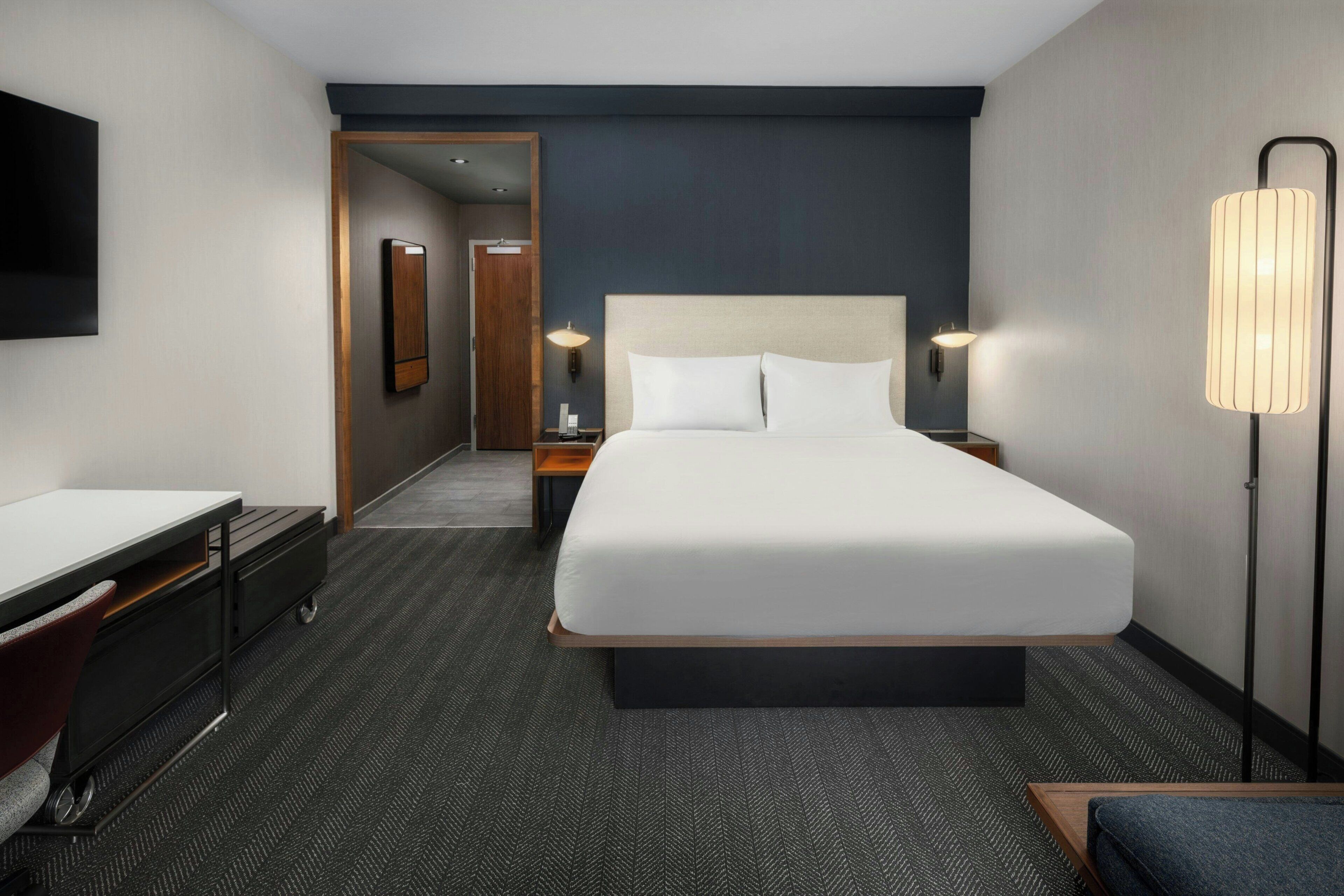 Habitación de hotel con una cama grande, un escritorio, una silla, una lámpara y un televisor.