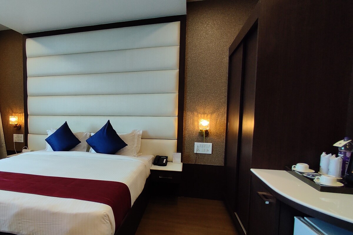 A hotel room with a large bed, two pillows, a bedside table with a phone, a chair, and a nightstand with a lamp.