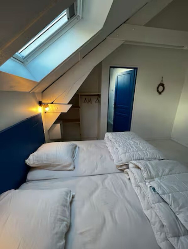 Una habitación pequeña y acogedora con techo inclinado, cama con ropa blanca, cabecera azul y puerta con marco azul.