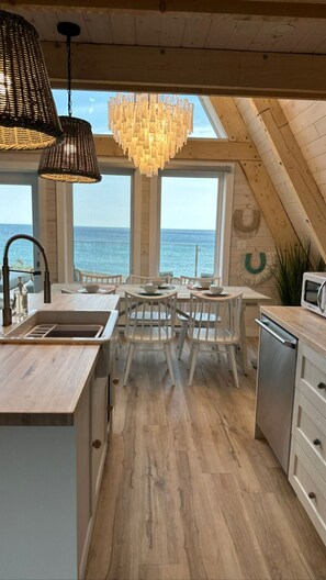 A kitchen with a view of the sea, wooden flooring, white cabinets, and a chandelier. - Mystical Beachfront Chalet , Hot tub , Sauna and Heated Container Pool (Cape Breton)