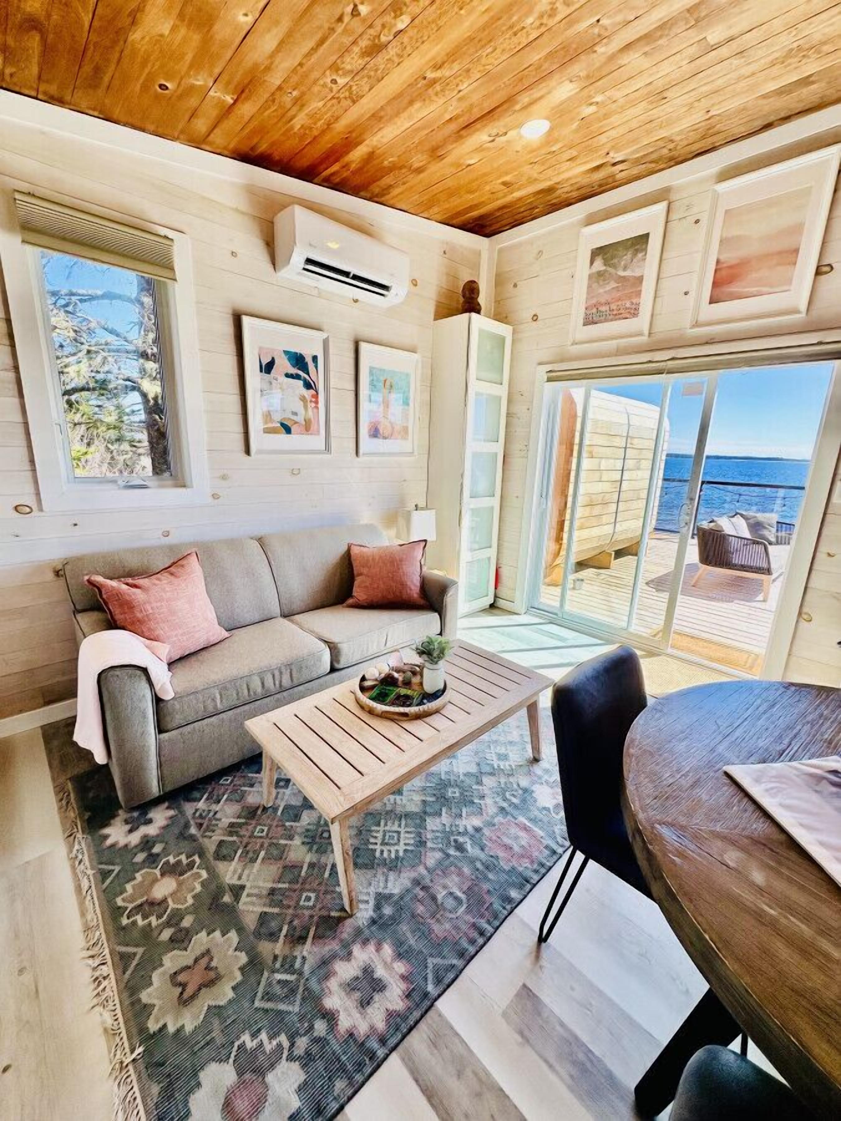 A cozy living room with a wooden ceiling, a grey sofa with cushions, a coffee table with a plant, and a view of the sea through large glass doors.
