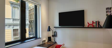 Ein Hotelzimmer mit Bett, Schreibtisch, Sessel, einem an der Wand befestigten Fernseher und einem Fenster mit Blick auf Gebäude.