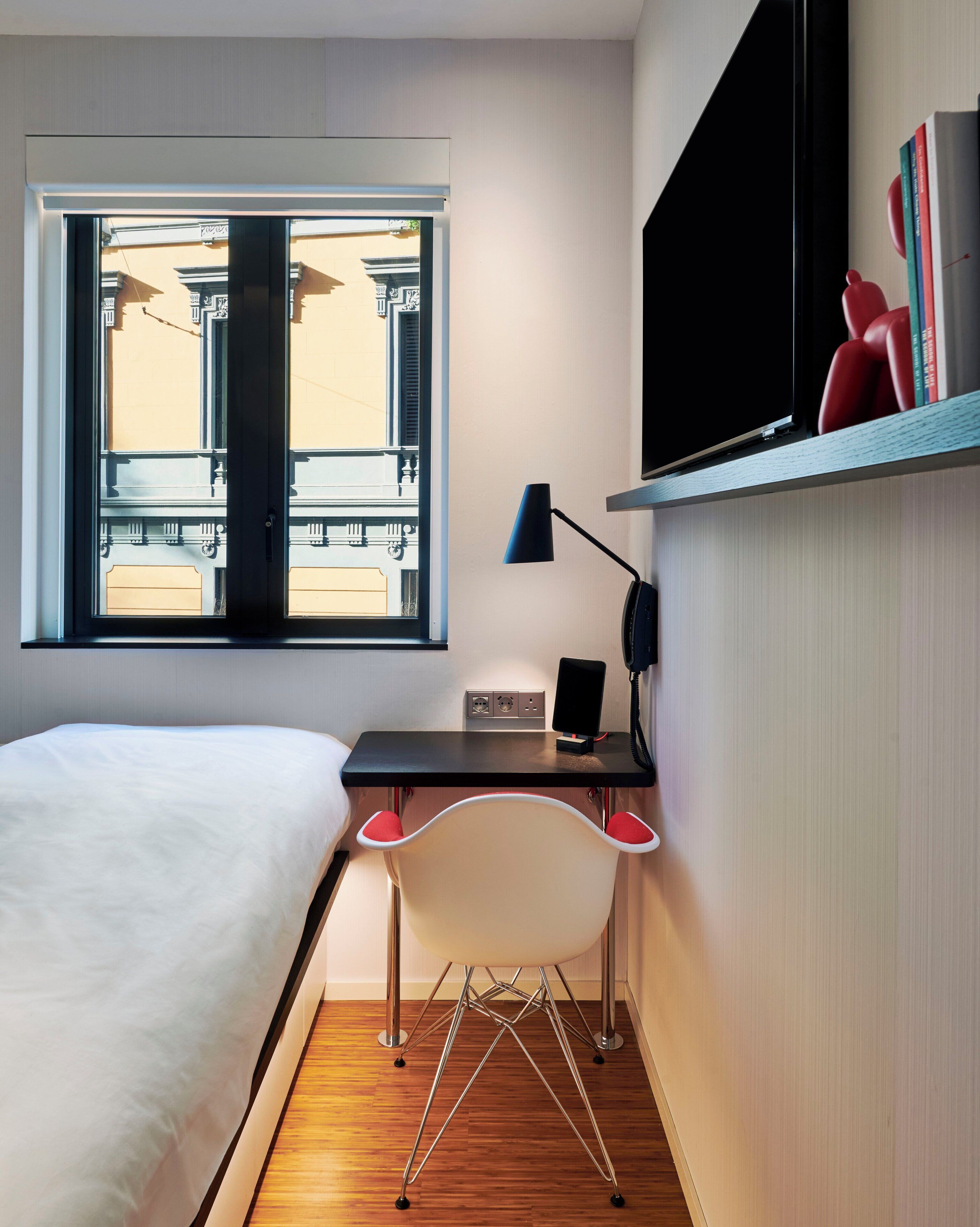 Una camera d'hotel con un letto, una scrivania con una sedia, una finestra e uno scaffale con libri.