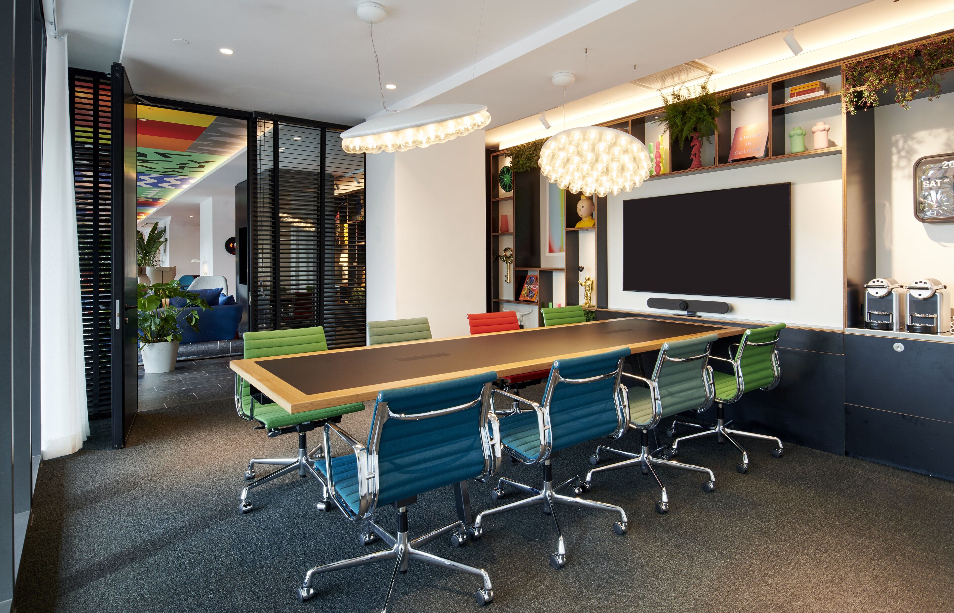 A modern conference room with a long table, green and blue chairs, a large flat-screen TV, and a decorative ceiling light.