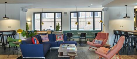 A modern living room with a blue sofa, a pink armchair, and a wooden coffee table. There are multiple seating options including a bench and chairs. The room features large windows with blinds and a view of the street outside.