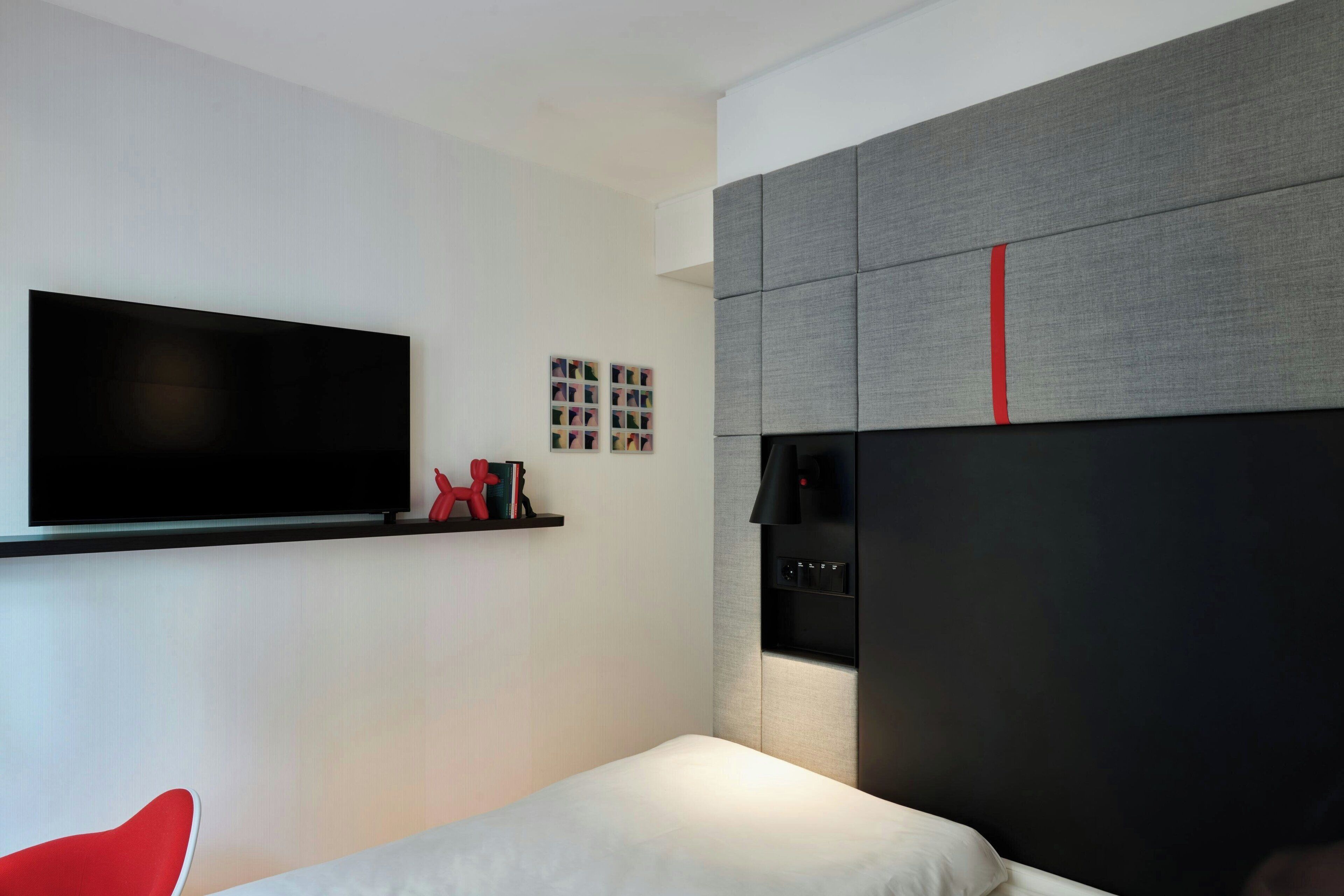 A modern hotel room with a large flat-screen TV mounted on the wall, a bed with a white mattress, a red chair, and a grey upholstered headboard with a red accent.