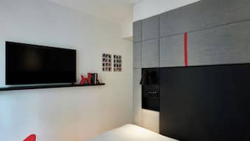 A modern hotel room with a large flat-screen TV mounted on the wall, a bed with a white mattress, a red chair, and a grey upholstered headboard with a red accent.