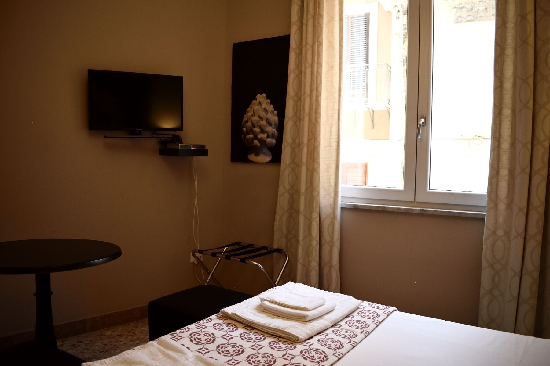 Quarto de hotel com cama, televisão na parede, mesa pequena e janela com cortinas.