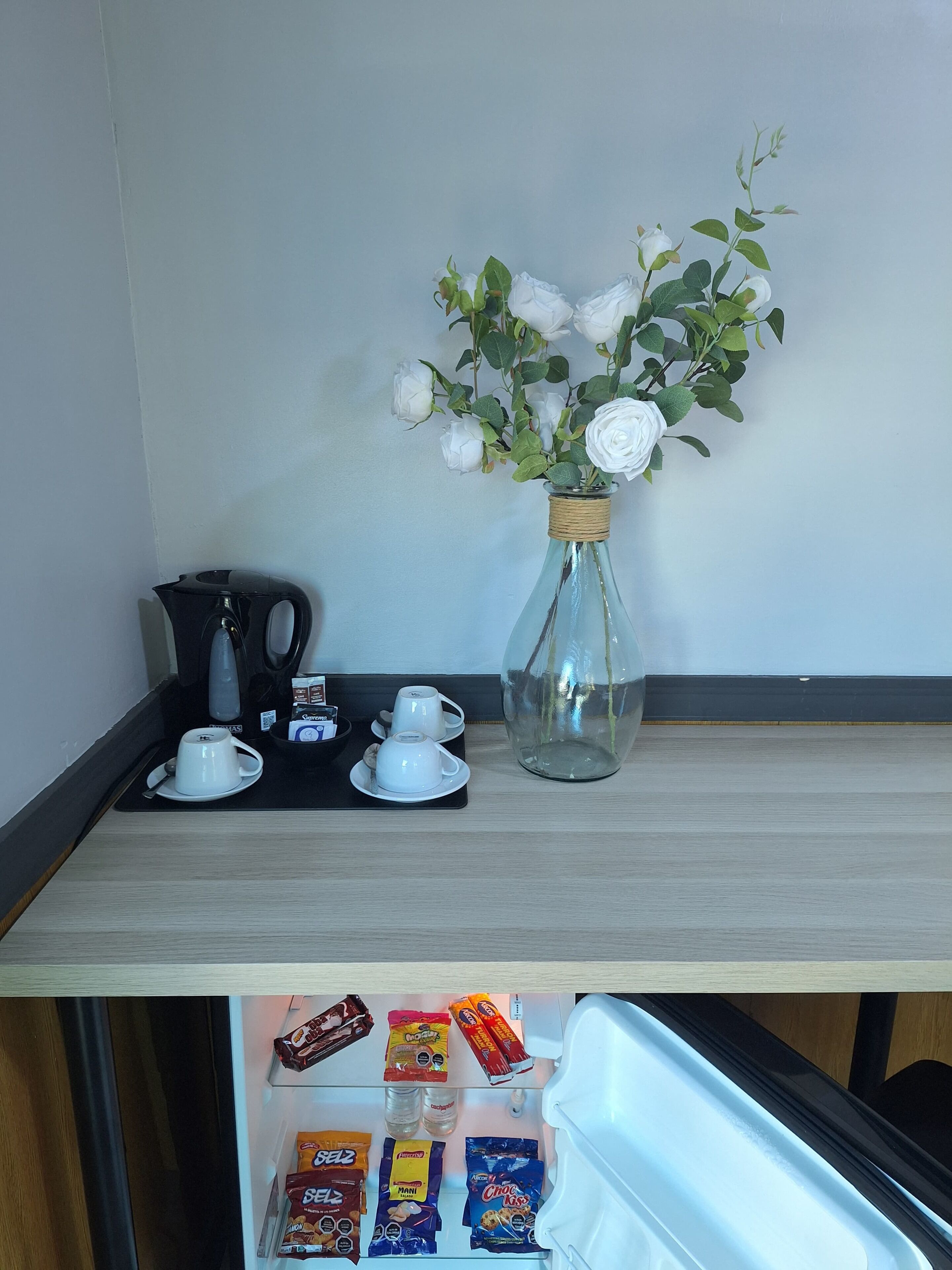 Una estación de café con una cafetera, dos tazas blancas y un florero con rosas blancas. Abajo, hay aperitivos y un refrigerador.