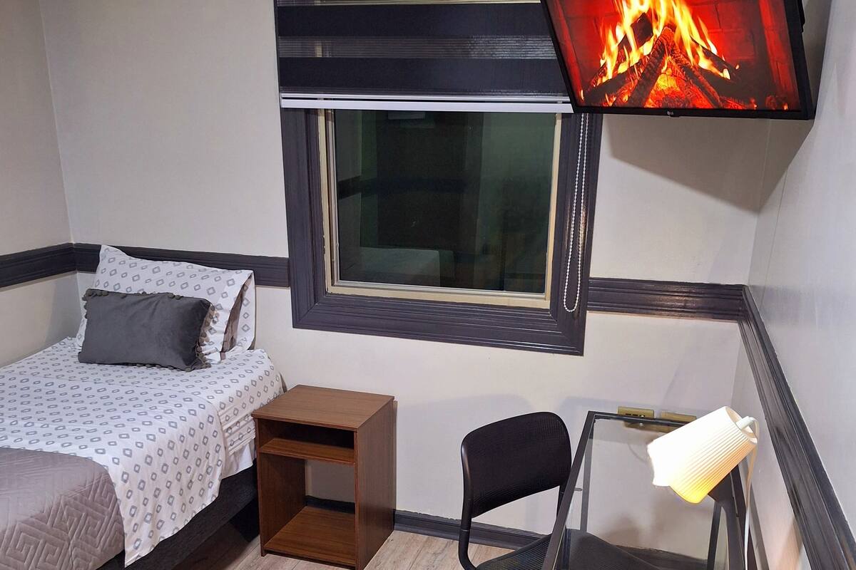 A compact room with a bed, a small desk with a chair, a TV mounted above the bed displaying a fireplace, and a window.
