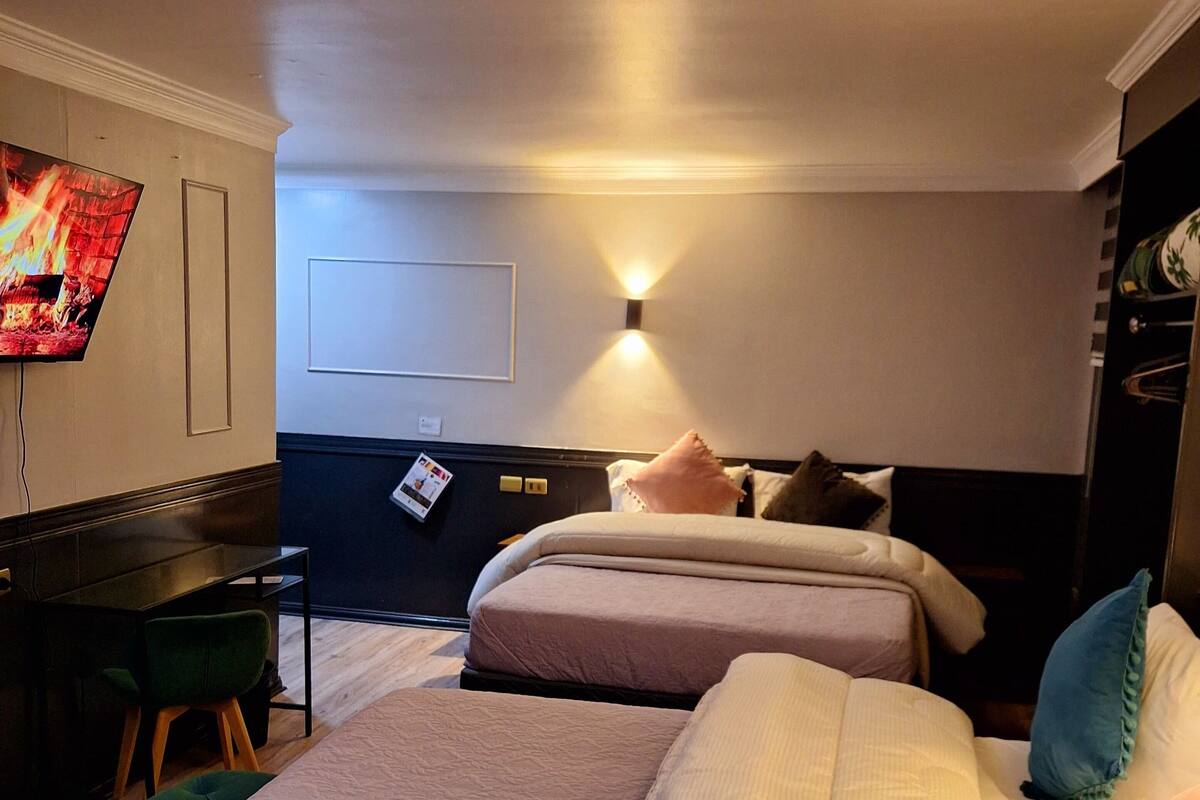 A hotel room with two beds, a desk, a chair, and a TV displaying a fireplace.