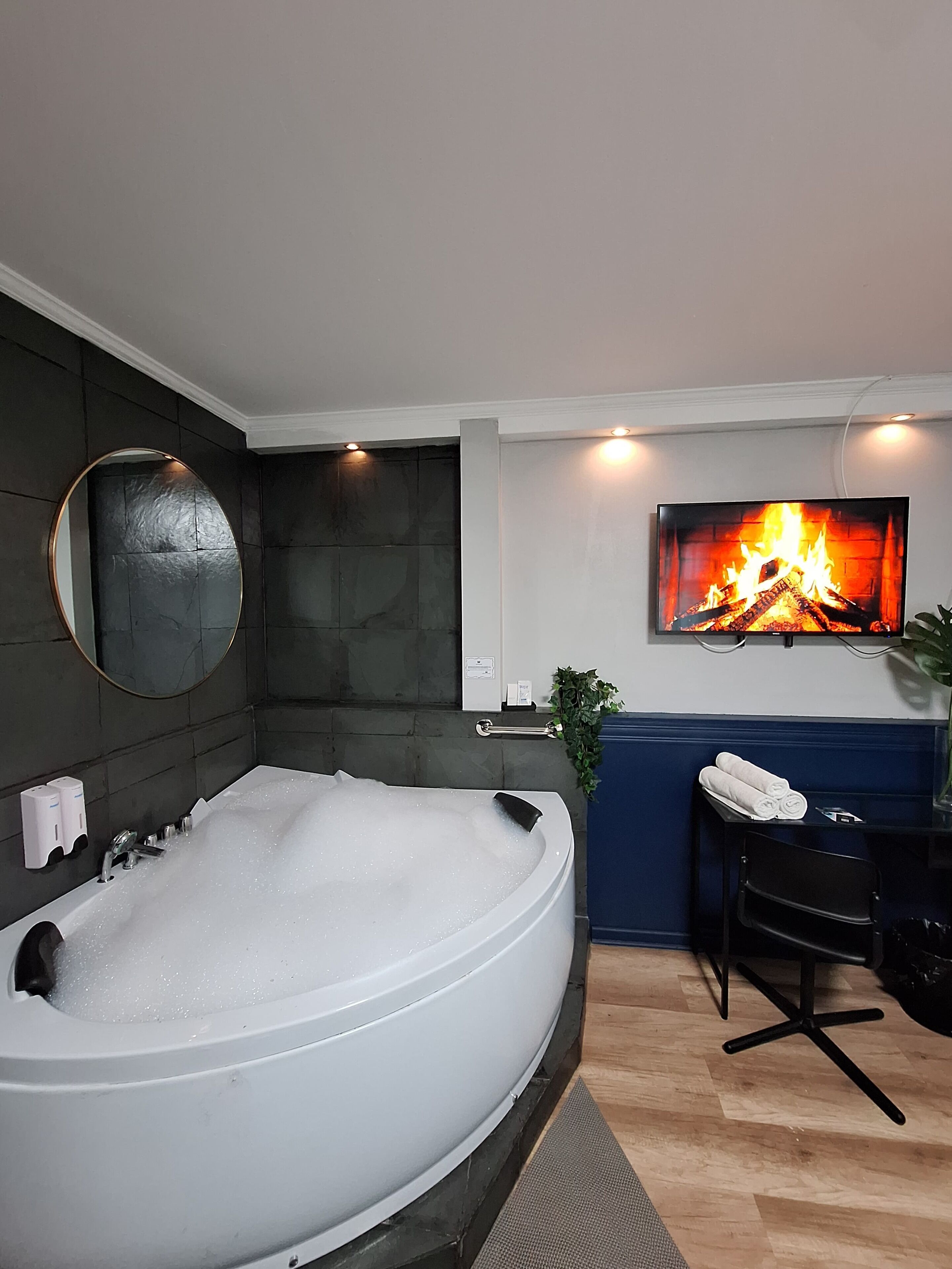 A modern bathroom with a large bathtub, a mirror, a wall-mounted TV displaying a fireplace, and a small table with towels.