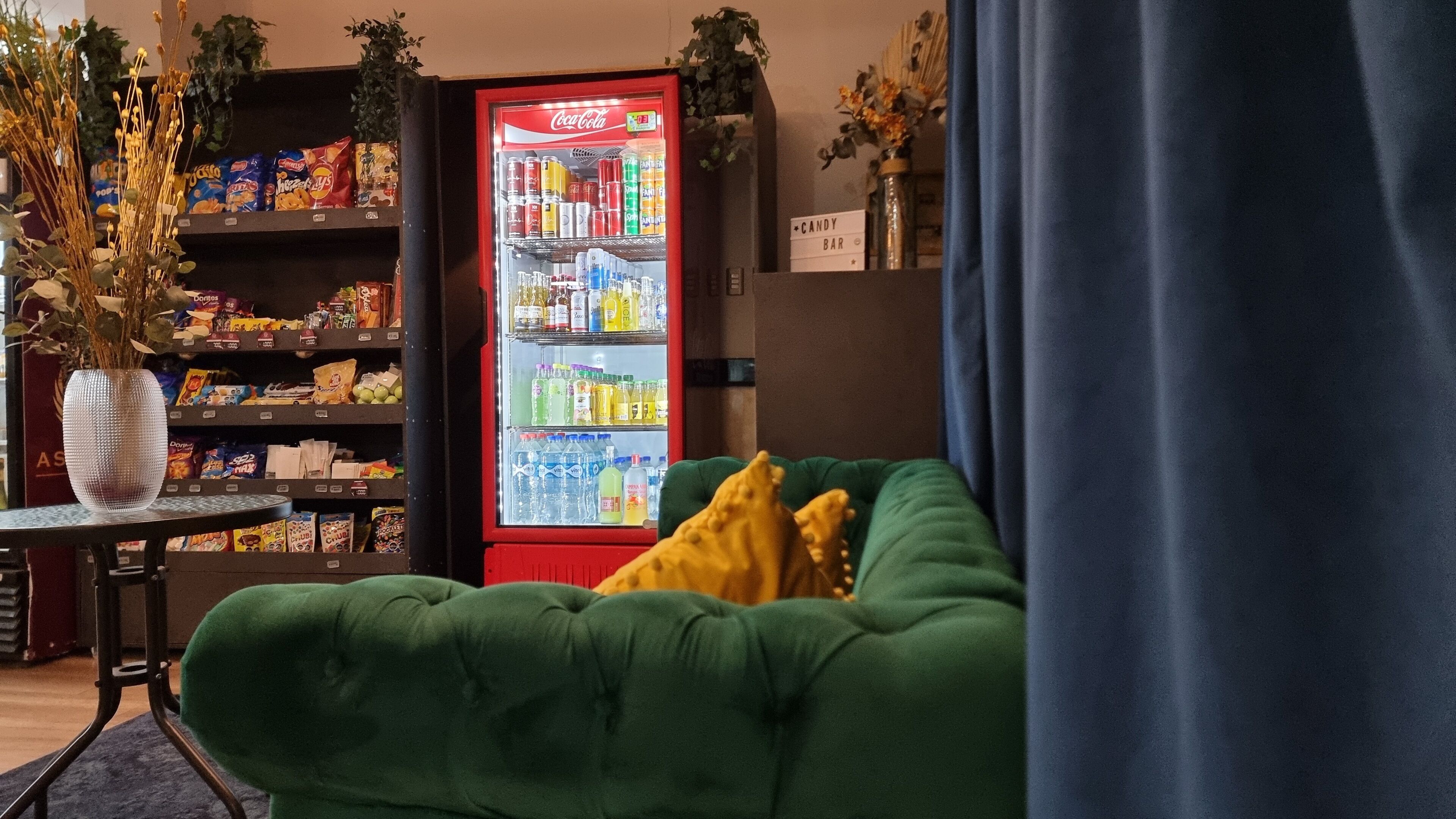 A room with a green sofa, a Coca-Cola refrigerator stocked with beverages, snack shelves, and a decorative vase.