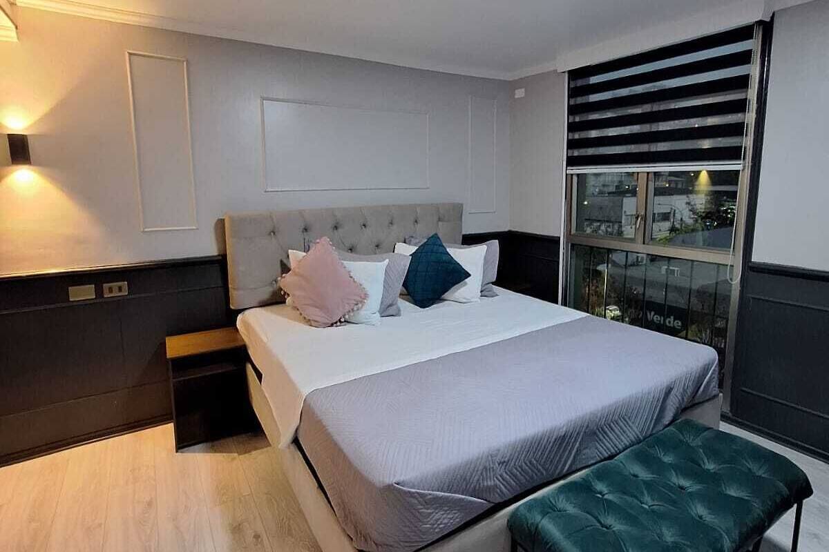 A modern bedroom with a large bed, bedside tables, a bench, and a window with blinds.