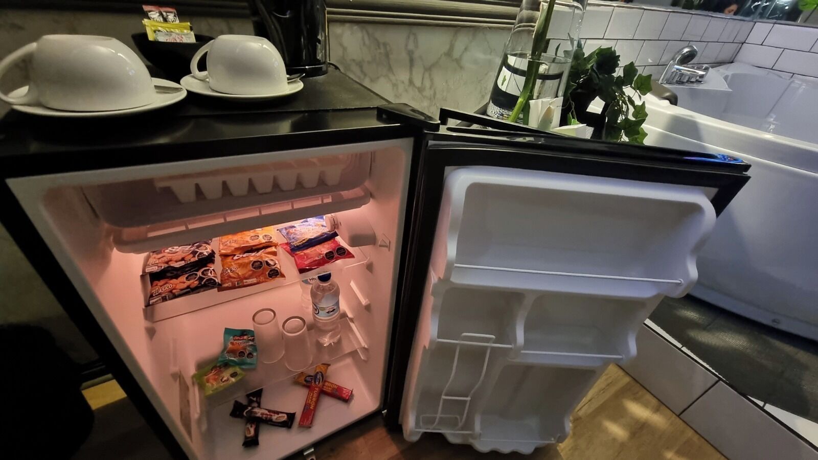 A refrigerator stocked with snacks and beverages, a sink, and a mirror in a kitchen setting.