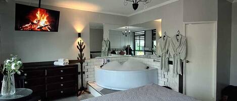 A modern hotel room with a large bed, a round jacuzzi, a TV mounted on the wall, and a mirror reflecting the room.