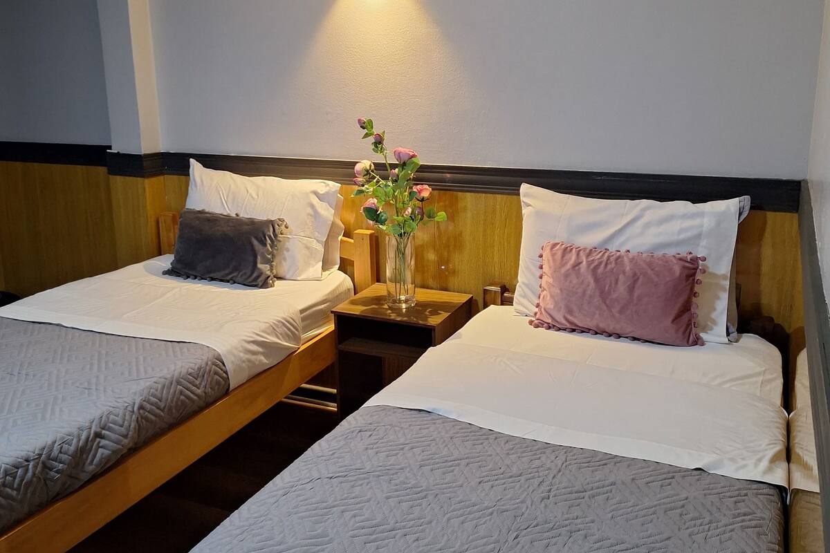 A hotel room with two single beds, a nightstand with a vase of flowers, and a wall-mounted light.