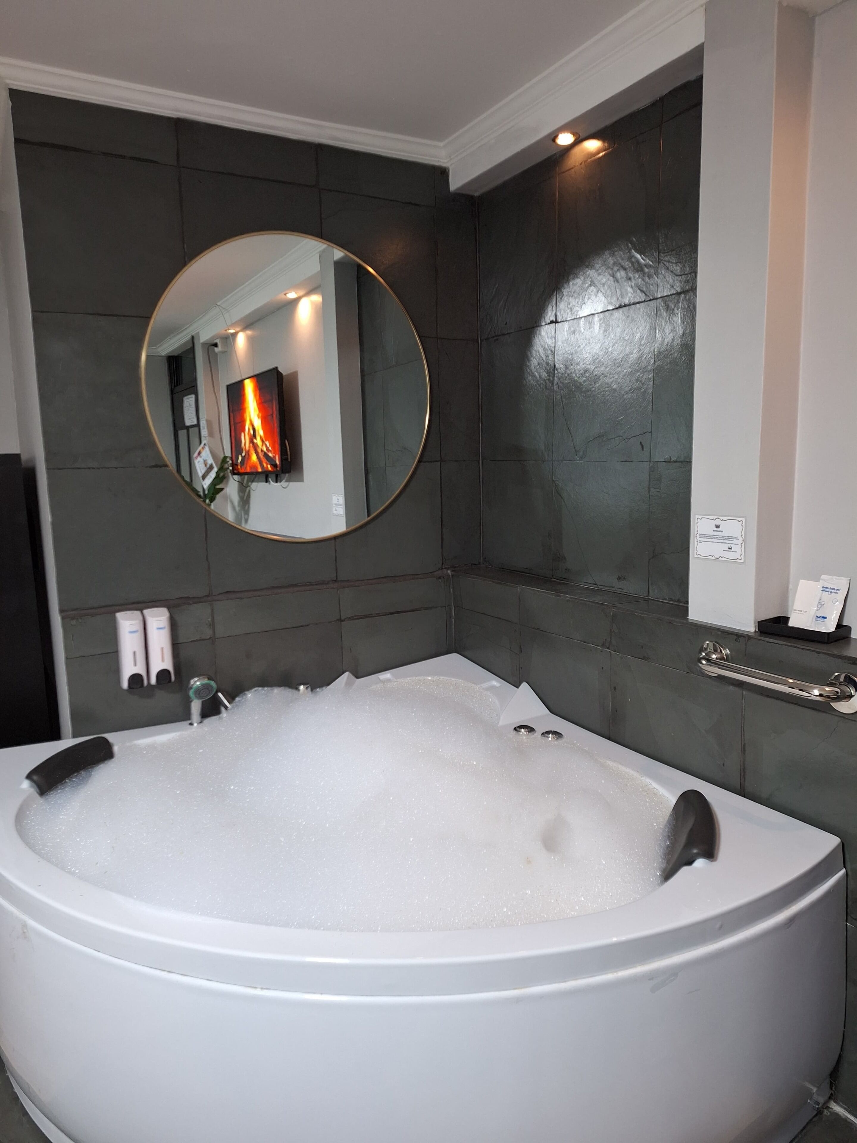 A modern bathroom with a large freestanding bathtub filled with bubbles, a round mirror, and a wall-mounted heater.