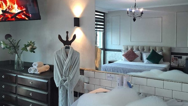 A modern bedroom with a large bed, a TV displaying a fire, a robe on a dresser, and a bathtub filled with bubbles. - Hotel Asturias (Vina del Mar)