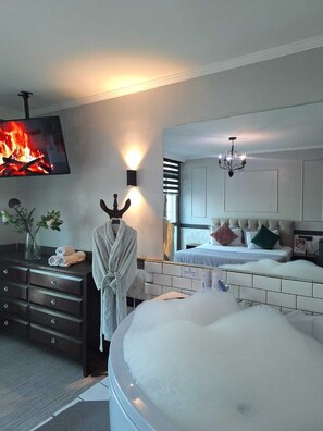 Un dormitorio moderno con una cama grande, un televisor mostrando un fuego, una bata en un tocador y una bañera llena de burbujas.