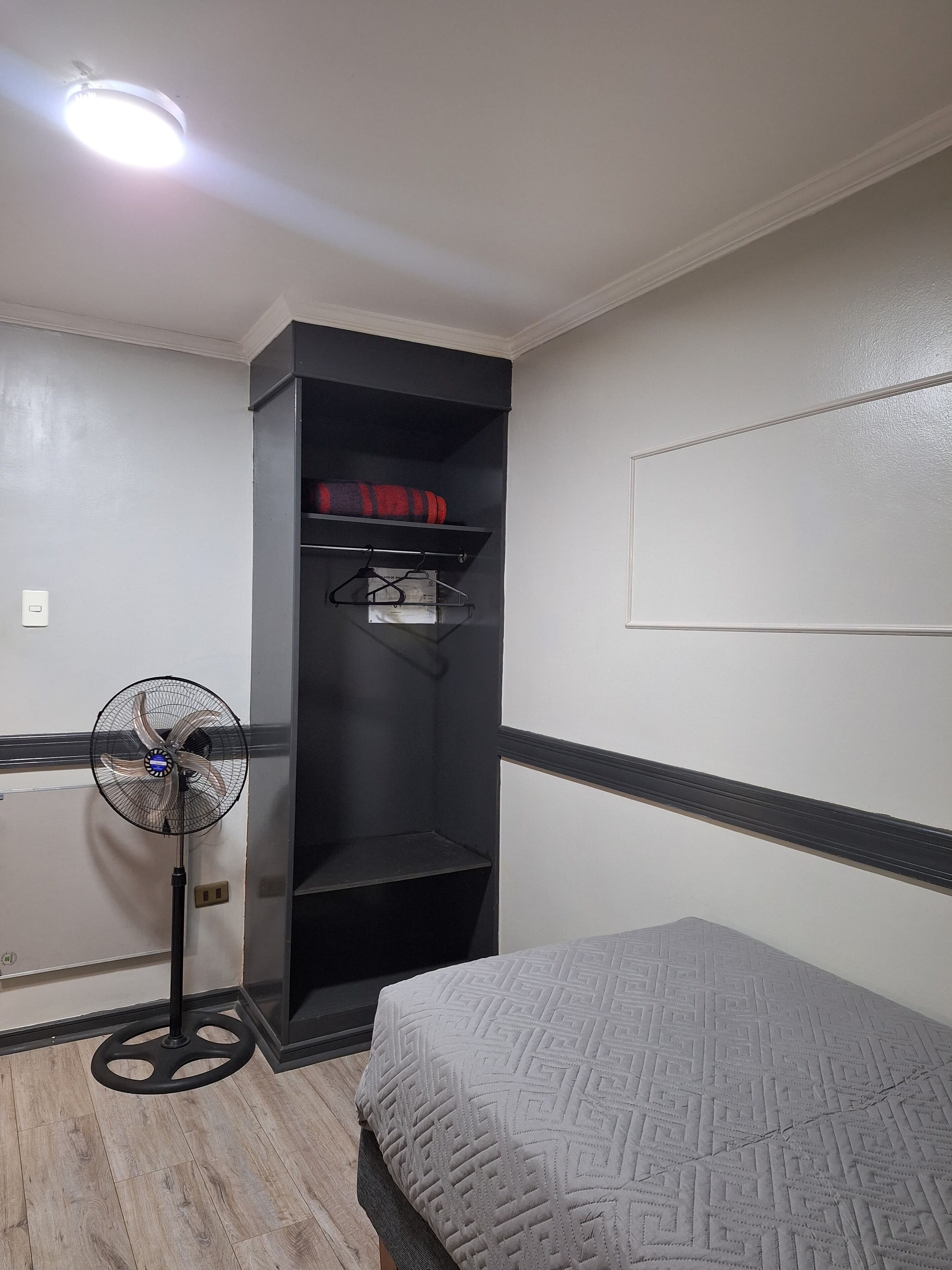 A room with a bed, a standing fan, a black shelving unit with clothes, and a light fixture.