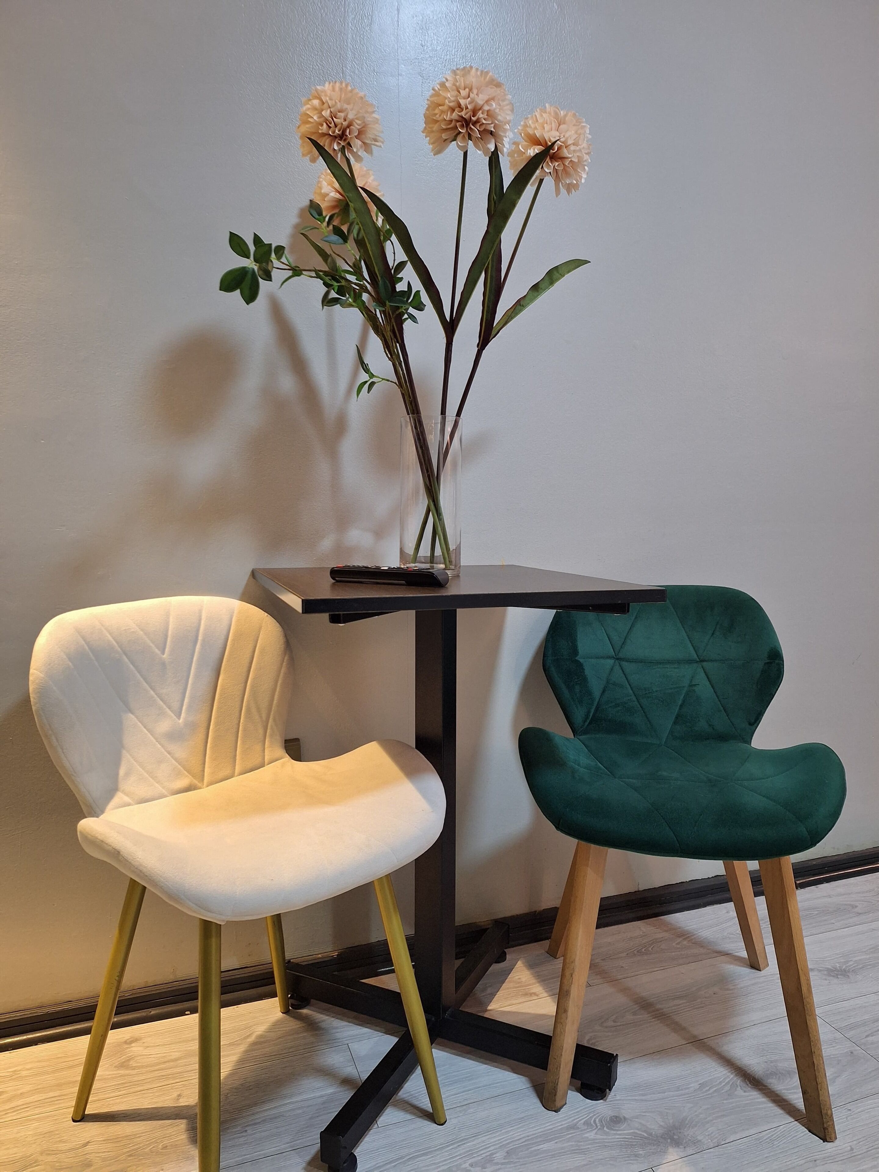 A room with a white and green upholstered chair, a small table with a vase of flowers, and a plain wall.