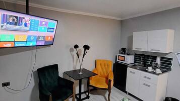 A compact living space with a television mounted on the wall, a small dining table, two chairs, a kitchen area with a microwave and a coffee maker, and a dark rug on the floor.