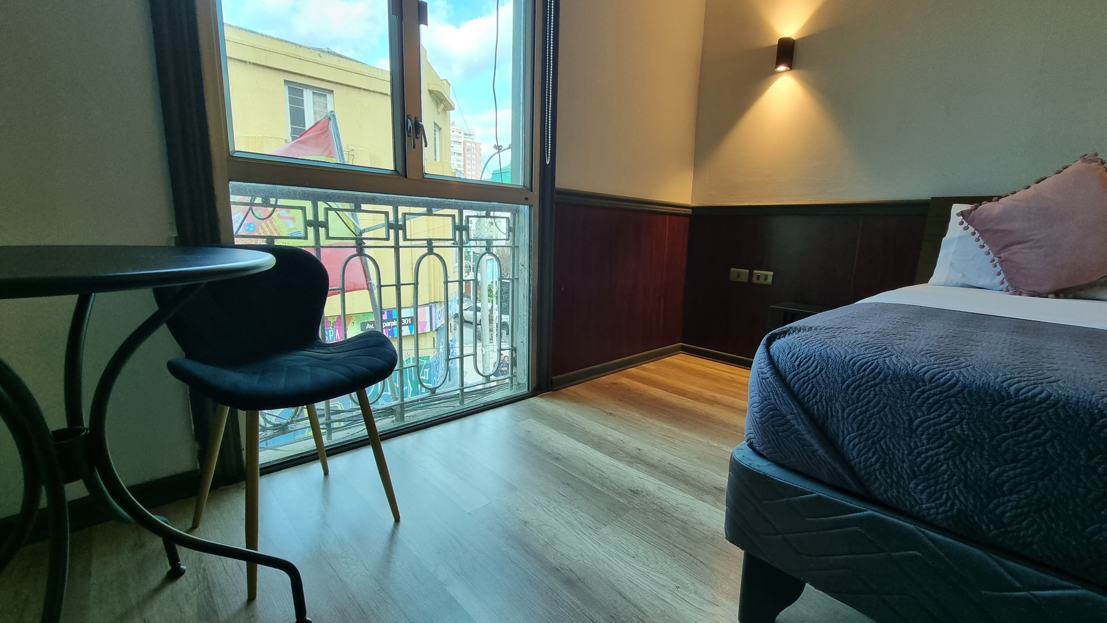 A hotel room with a bed, a small table, a chair, and a window with a view of buildings and a balcony.