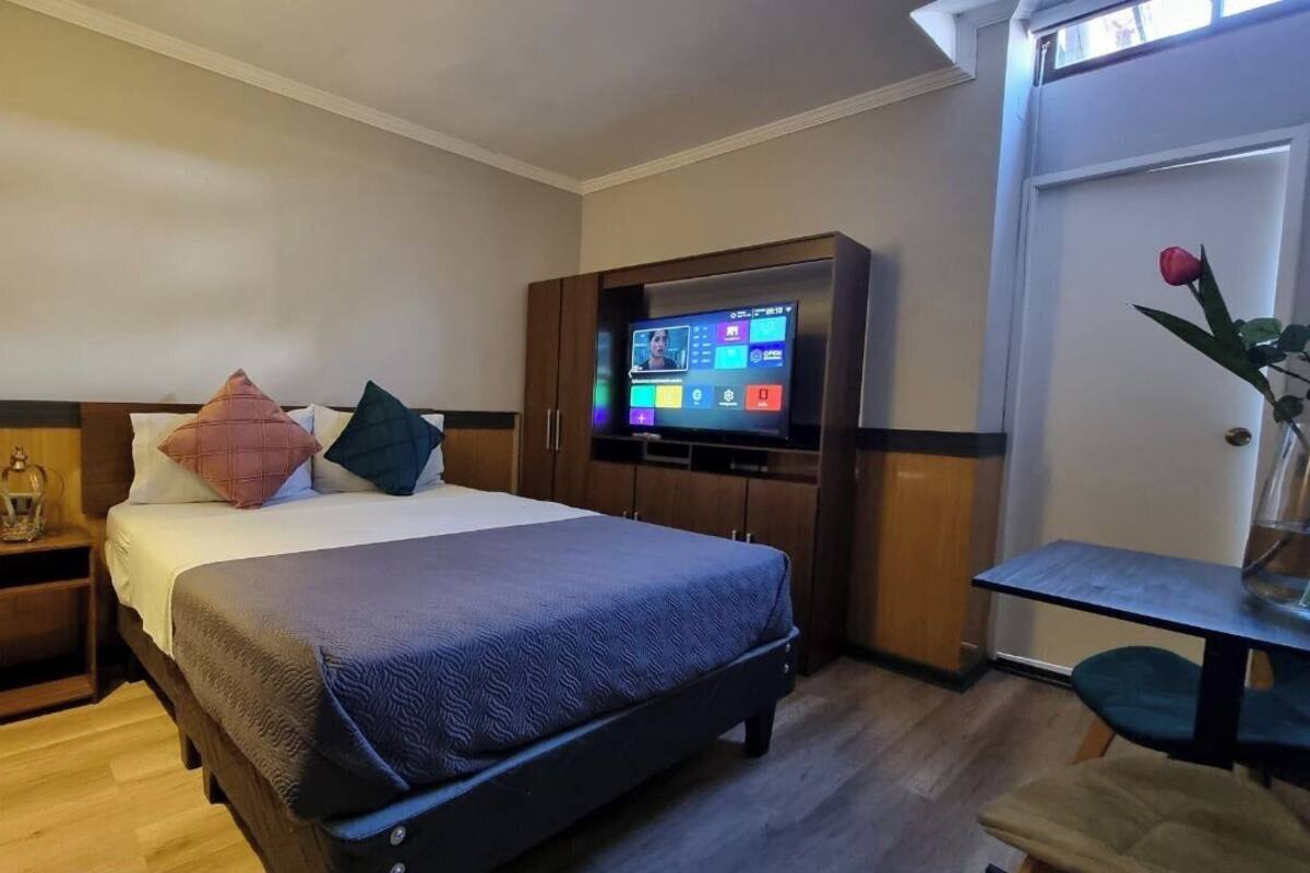 A hotel room with a bed, a TV mounted on the wall, a small table, and a vase with a flower.