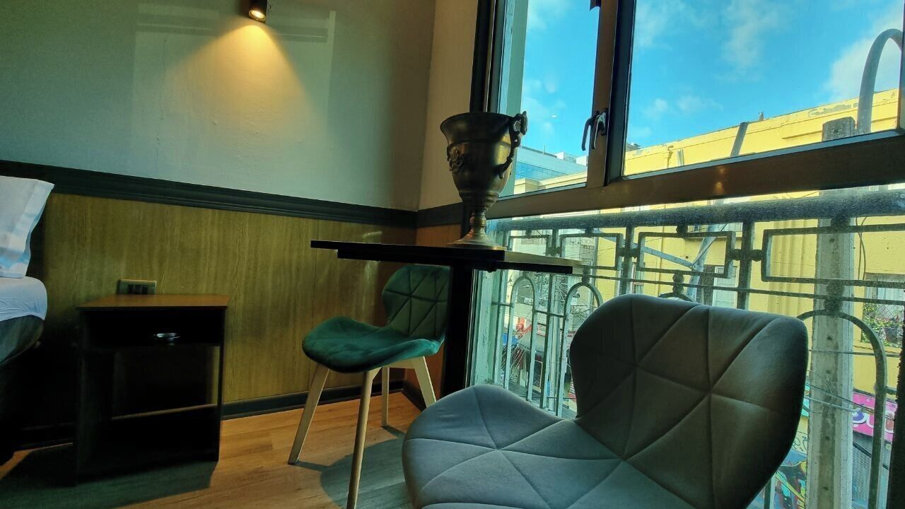 Habitación de hotel con una cama, una silla verde, una mesita con un trofeo y una ventana que da a edificios y un balcón.