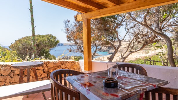 Un espacio de estar al aire libre cubierto, con una mesa y sillas, que ofrece vistas panorámicas del mar y árboles.