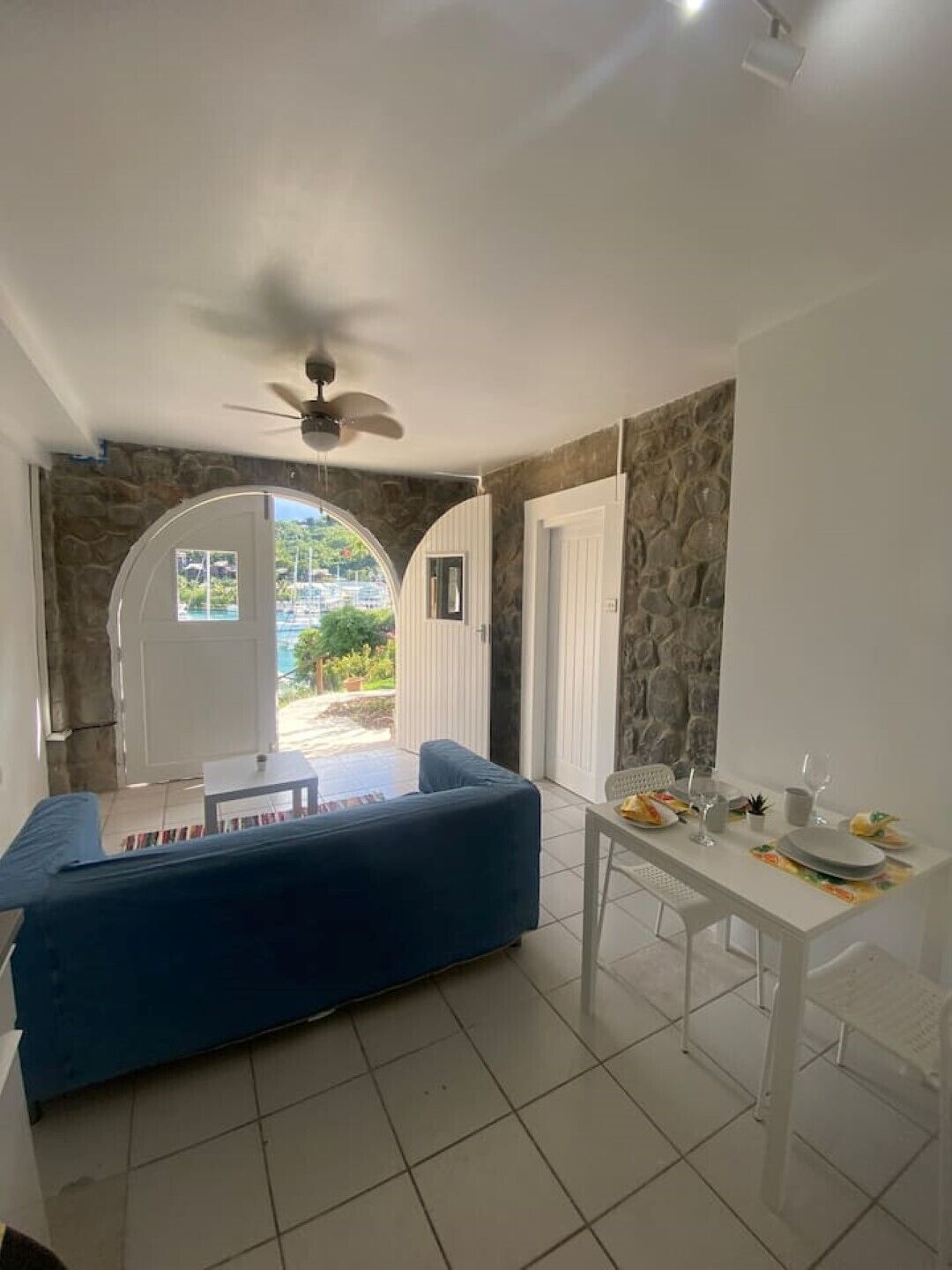 A room with a blue sofa, a dining table set with plates and glasses, a ceiling fan, and a view of a pool and greenery through an arched doorway.