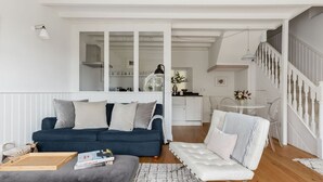 A modern living room with a dark blue sofa, a white armchair, and a grey ottoman. There is a wooden coffee table with books and a tray. A kitchen is visible through a glass door, and a staircase leads to an upper level. - Beautiful converted stable near the coast (Plymouth)