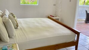 Ein Schlafzimmer mit einem hölzernen Bett, einem Deckenventilator, einer Klimaanlage, einem Fenster mit einem floralen Vorhang und Blick auf Grünfläche.