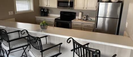 A kitchen with white cabinets, a stainless steel refrigerator, a microwave, a stove, and two bar stools with cushioned seats.