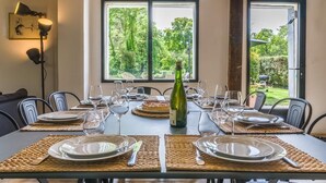 Mesa de comedor preparada para seis personas, con posavasos, platos, cubiertos y copas de vino. Una botella de vino está colocada en el centro. La habitación tiene vistas a zonas verdes a través de amplios ventanales.